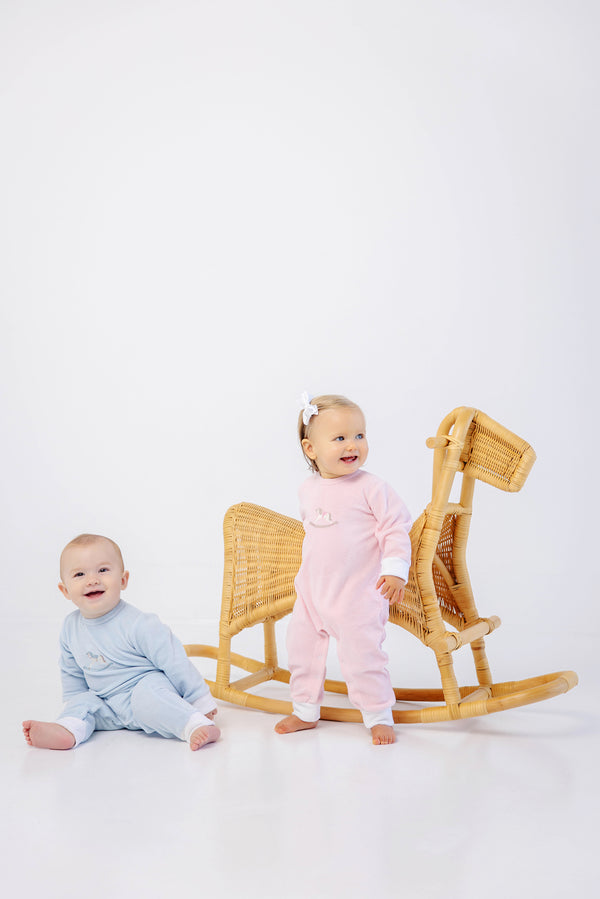 Two babies in pastel pajamas pose near a wicker rocking horse. One baby sits on the floor in a blue playsuit, while the other stands beside the rocking horse wearing The Beaufort Bonnet Company’s Paxton Playsuit in Palm Beach Pink with Rocking Horse Embroidery and a white bow headband. The background is white and minimal.