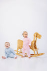 Two babies in pastel outfits, one sitting in The Beaufort Bonnet Company’s Paxton Playsuit - Buckhead Blue with Rocking Horse Embroidery and one standing in pink, are posing beside a wicker rocking horse against a white background.