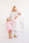 A woman in a white lace dress holds a baby in a Cameron Cross Front Set - Palm Beach Pink with Hamptons Hot Pink Little Sister, while a young girl in a pink gingham top and pink shorts stands beside her.