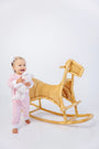 A smiling baby wearing The Beaufort Bonnet Company’s Paxton Playsuit in Palm Beach Pink with Rocking Horse Embroidery holds a stuffed animal while standing next to a wicker rocking horse against a white background.