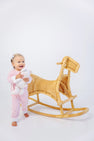 A smiling baby wearing The Beaufort Bonnet Company’s Paxton Playsuit in Palm Beach Pink with Rocking Horse Embroidery holds a stuffed animal while standing next to a wicker rocking horse against a white background.