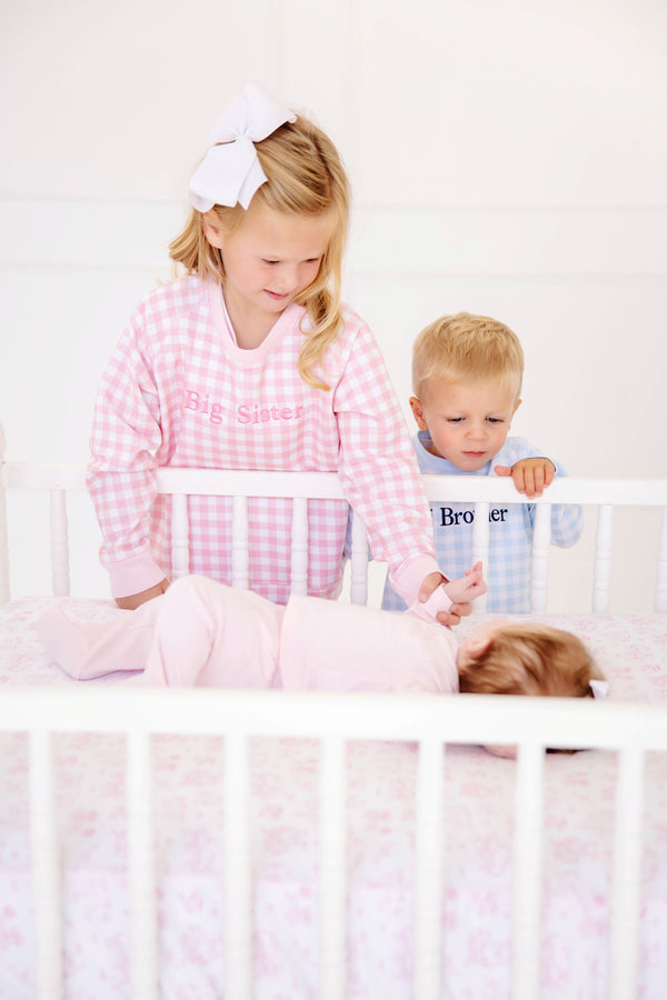 A young girl in a pink gingham sibling reveal shirt and a boy in a Cassidy Comfy Crewneck - Buckhead Blue Gingham with Nantucket Navy Big Brother look down at a baby lying in a white crib.