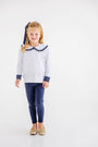 A young child with blonde hair stands and smiles against a plain white background, wearing The Beaufort Bonnet Company’s Maude’s Ruffle Cassidy Comfy Crewneck in Nantucket Navy Microdot with Worth Avenue White. She pairs the top with navy leggings, gold shoes, and a matching navy bow in her hair.