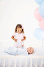 A young girl in a pink dress looks down into a crib at a baby wearing a Cameron Cross Front Set - Buckhead Blue with Nantucket Navy Little Brother, accompanied by blue and pink balloons for this baby announcement.