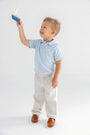 A young boy in The Beaufort Bonnet Company’s Prim & Proper Polo - Buckhead Blue with Bellport Butter Yellow Stork and beige pants smiles while holding up a small toy sailboat.