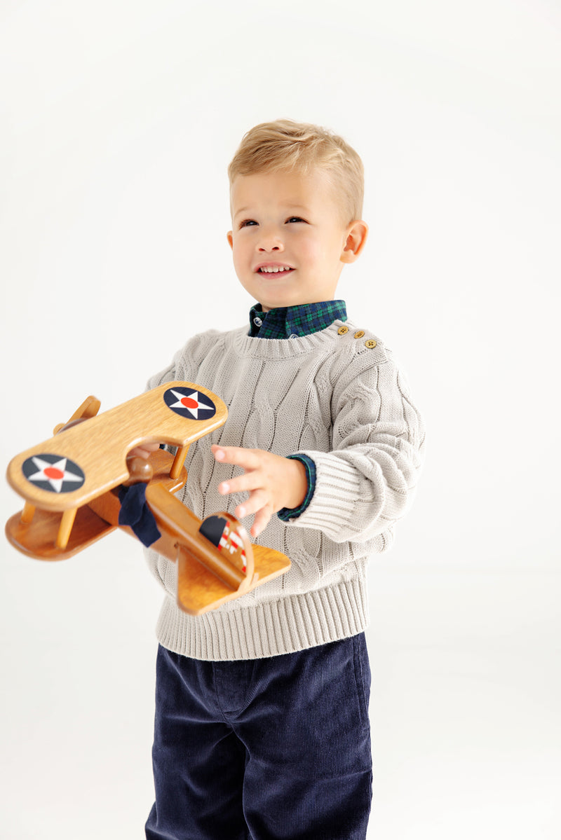 A young boy in a cream sweater and dark pants smiles while holding a wooden toy airplane with star decals, standing against a plain white background, with The Beaufort Bonnet Company’s Dean’s List Dress Shirt in Woodlake Point Plaid peeking out at the collar.
