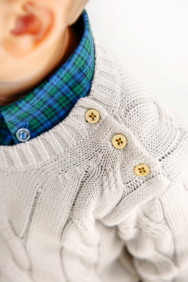 Close-up of a child wearing a cream cable-knit sweater with three tan buttons on the shoulder, layered over The Beaufort Bonnet Company’s Dean’s List Dress Shirt in Woodlake Point Plaid. Only the lower face, ear, and shoulder are visible.