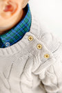 Close-up of a child wearing a cream cable-knit sweater with three tan buttons on the shoulder, layered over The Beaufort Bonnet Company’s Dean’s List Dress Shirt in Woodlake Point Plaid. Only the lower face, ear, and shoulder are visible.