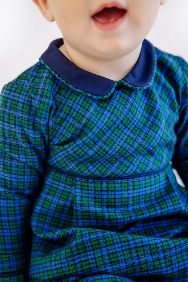 A young child wearing The Beaufort Bonnet Company’s Long Sleeve Bradford Romper in Woodlake Point Plaid with Nantucket Navy, smiling with their mouth slightly open. The image is cropped to show only the child’s lower face and upper body.
