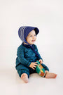 A smiling baby in a blue bonnet and The Beaufort Bonnet Company’s Long Sleeve Bradford Romper – Woodlake Point Plaid with Nantucket Navy sits on the floor, holding a colorful toy bus.