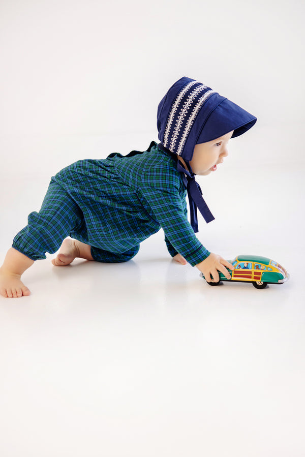 A baby in a blue bonnet and The Beaufort Bonnet Company Long Sleeve Bradford Romper in Woodlake Point Plaid with Nantucket Navy crawls on the floor, pushing a colorful toy bus.