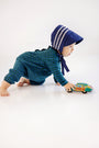 A baby in a blue bonnet and The Beaufort Bonnet Company Long Sleeve Bradford Romper in Woodlake Point Plaid with Nantucket Navy crawls on the floor, pushing a colorful toy bus.