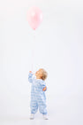 A toddler wearing The Beaufort Bonnet Company Long Sleeve Mary Dal Romper in Blue Choo Choos stands on a white background, looking up and holding a pink balloon by its string.