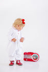 A toddler in a Long Sleeve Britt Romper - Worth Avenue White with Holly stands looking down at a vintage-style red radio on the floor.