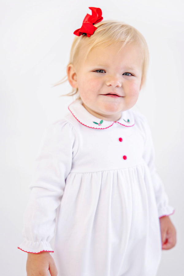 A toddler wearing The Beaufort Bonnet Company Long Sleeve Britt Romper - Worth Avenue White with Holly, a white Pima cotton outfit with a Peter Pan collar featuring embroidered holly, red trim, and a matching red hair bow.
