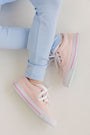 A child wearing The Beaufort Bonnet Company’s Mitzy Sue Slacks - Beale Street Blue and pale pink sneakers with white laces sits cross-legged on a white surface.