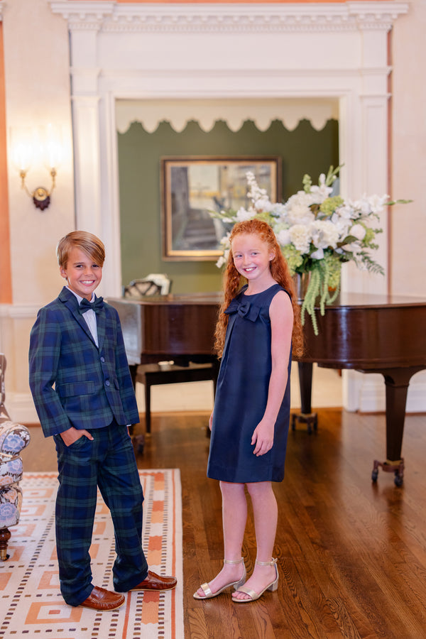 Two smiling children, a boy wearing The Beaufort Bonnet Company’s Baylor Bowtie - Berwick Black Watch plaid suit and bow tie and a girl in a navy dress and sandals, stand on a wooden floor in an elegant room with a grand floral arrangement and a piano in the background.