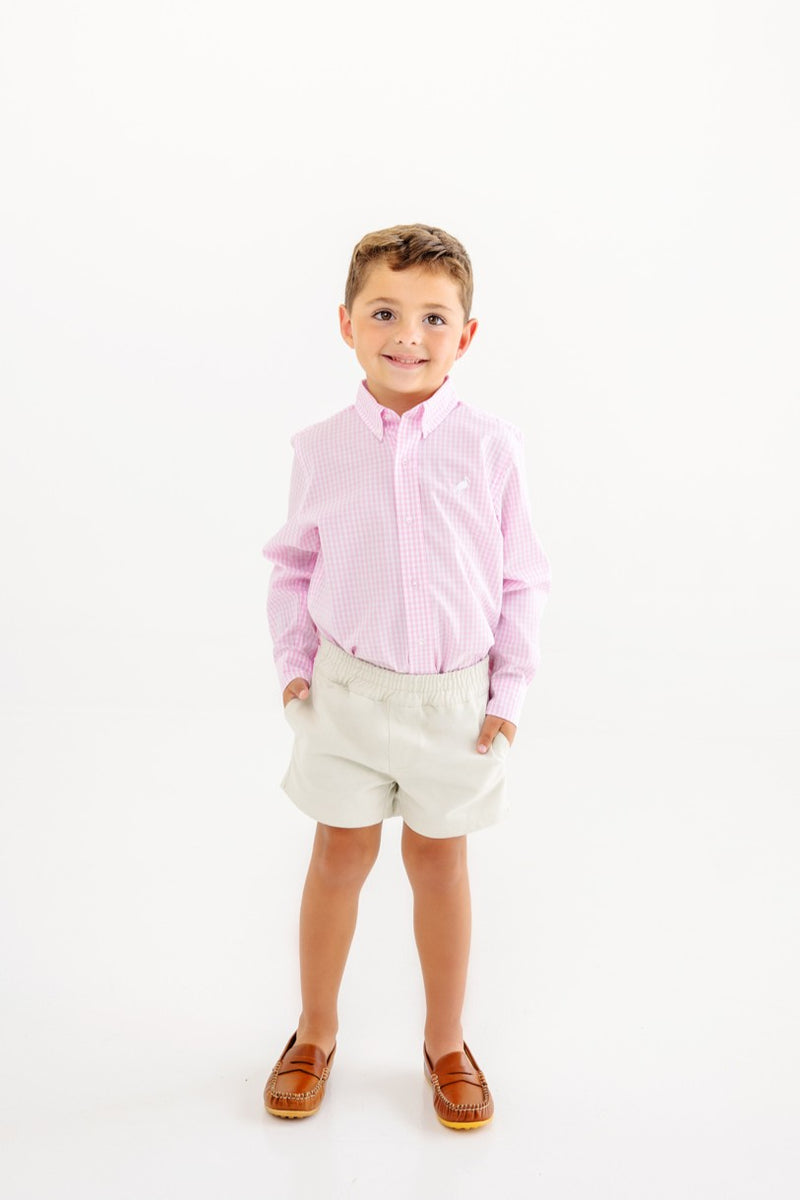A young boy stands smiling with his hands in his pockets, wearing The Beaufort Bonnet Company’s Dean’s List Dress Shirt in the Pink Greenbrier Gingham with Worth Avenue White Stork, paired with beige shorts and brown loafers against a plain white background.