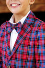 A smiling child wears The Beaufort Bonnet Company’s Grandccasion™ Booker Blazer – Bucktown Tartan with Barrington Blue, paired with a matching bow tie and white shirt. Only the lower half of the face and torso are visible.
