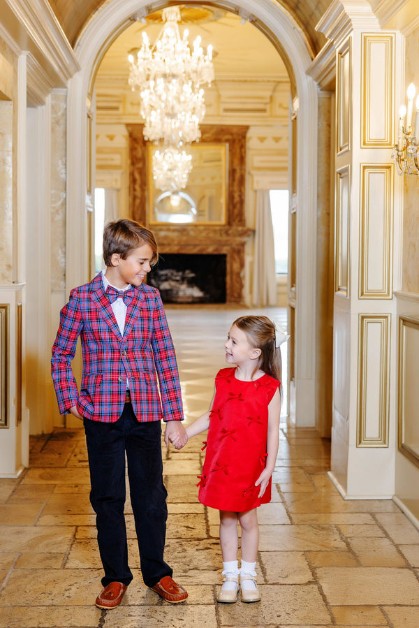 A boy dressed in The Beaufort Bonnet Company’s Prep School Pants – Newport Night Corduroy with Richmond Red Stork, a plaid blazer, and a bow tie holds hands with a girl in a red dress. They smile at each other in an elegant hallway adorned with chandeliers and ornate decor.