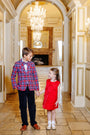 A boy dressed in The Beaufort Bonnet Company’s Prep School Pants – Newport Night Corduroy with Richmond Red Stork, a plaid blazer, and a bow tie holds hands with a girl in a red dress. They smile at each other in an elegant hallway adorned with chandeliers and ornate decor.