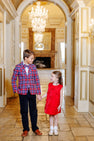 A boy dressed in The Beaufort Bonnet Company’s Prep School Pants – Newport Night Corduroy with Richmond Red Stork, a plaid blazer, and a bow tie holds hands with a girl in a red dress. They smile at each other in an elegant hallway adorned with chandeliers and ornate decor.