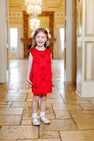 A young girl stands smiling on a tiled floor in an elegant, ornate room with chandeliers and gold accents, wearing The Beaufort Bonnet Company’s Grandccasion™ Jilly Jumper in Richmond Red Taffeta adorned with bows.