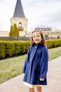 A young girl wearing The Beaufort Bonnet Company’s Penelope Peacoat in Nantucket Navy stands smiling on a path, with manicured hedges and a stone building featuring a tall pointed tower in the background.