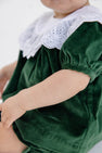 A baby wearing The Beaufort Bonnet Company’s Short Sleeve Bon Air Bloomer Set in Grier Green Velveteen with Worth Avenue White, featuring short puffy sleeves and a white lace collar, sits with one arm extended.