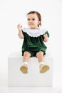 A toddler with a white bow in her hair wears The Beaufort Bonnet Company’s Short Sleeve Bon Air Bloomer Set in Grier Green Velveteen with Worth Avenue White, featuring a white lace collar, as she sits on a white block against a plain white background.