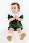 A toddler dressed in The Beaufort Bonnet Company’s Short Sleeve Bon Air Bloomer Set in Grier Green Velveteen with Worth Avenue White sits on a white block, looking up with hands clasped and legs dangling.