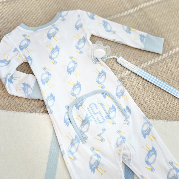 A long-sleeved blue heron print baby romper with a monogrammed front pocket, paired with a Parker Paci Clip - Blue Grand Gasparilla Gingham with Buckhead Blue on a blue and white checkered ribbon.