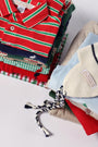 A neatly folded stack of brightly colored children’s clothes—including striped shirts, patterned sweaters such as The Beaufort Bonnet Company’s Hayden Half-Zip Sweater in Richmond Red with Nantucket Navy Stork, and a knit hat with navy and white braided ties—arranged on a white surface.