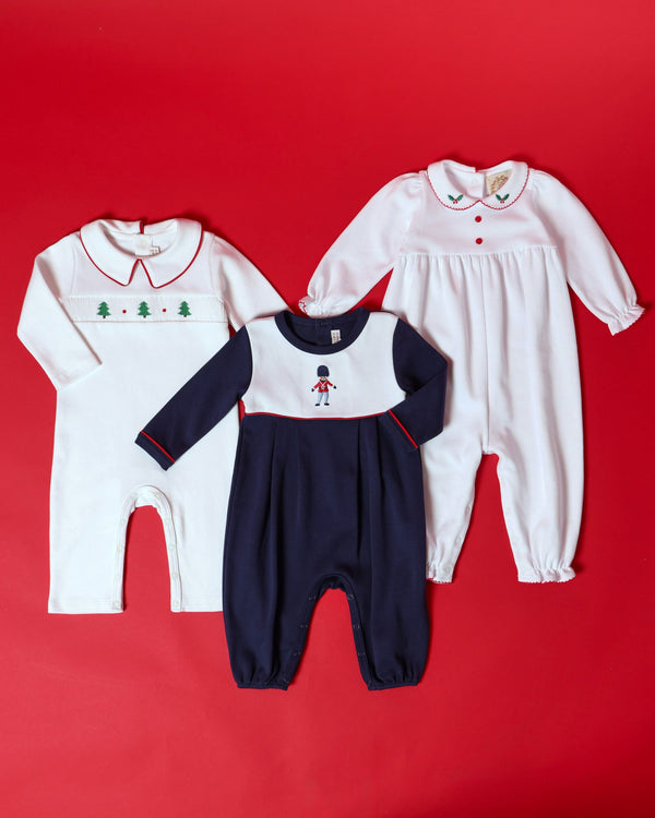 Three Pima cotton Christmas rompers: a Long Sleeve Britt Romper - Worth Avenue White with Holly, a white romper with a green tree pattern, and a navy and white toy soldier emblem romper.
