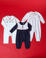 Three Pima cotton Christmas rompers: a Long Sleeve Britt Romper - Worth Avenue White with Holly, a white romper with a green tree pattern, and a navy and white toy soldier emblem romper.