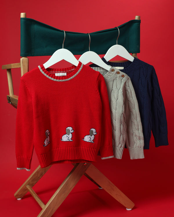 Three sweaters hanging on a director’s chair: a red Crawford Crewneck embroidered with dachshunds, a Crawford Crewneck - Grantley Gray with Brass Buttons cable-knit sweater, and a solid navy cable-knit sweater.