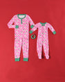 Two sets of matching “Noelle’s Night Night - Bows of Holly with Kiawah Kelly Green” pajamas from The Beaufort Bonnet Company—one for an adult and one for a baby—are displayed on a red background beside a small green and red wreath. Crafted from soft Pima cotton, these cozy sleepers feature festive pink fabric with green trim and holly bow patterns, offering comfort and holiday style for the whole family.