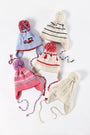 Five colorful winter hats with pom-poms and ear flaps are laid out on a white surface. Each hat showcases a unique pattern, featuring stripes, geometric designs, and festive motifs. Among them is the playful Parrish Pom Pom Hat - Palmetto Pearl with Bows & Trees from The Beaufort Bonnet Company, adding an extra touch of cheer to the collection.