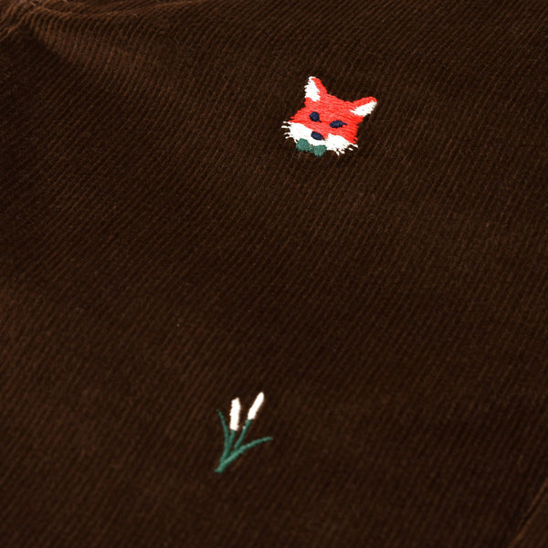 A close-up of The Beaufort Bonnet Company’s Critter Sheffield Pants in Chelsea Chocolate Corduroy, featuring a small embroidered red fox head and a tuft of green grass with two white tips.