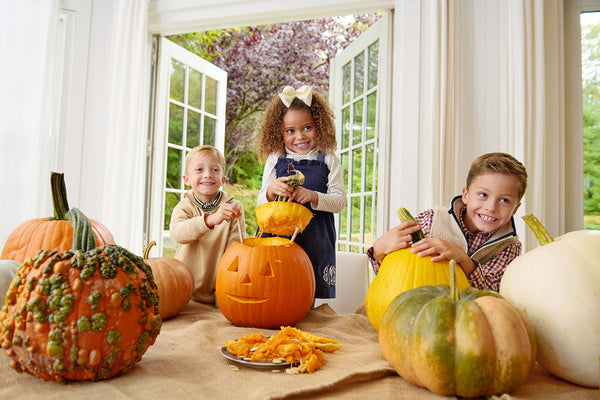 Three smiling children carve pumpkins indoors, surrounded by various pumpkins and pumpkin seeds on a table. Bright daylight streams in through open double doors to a lush green garden outside, while one child shows off their Macy Drew Dress - Nantucket Navy Corduroy from The Beaufort Bonnet Company—a charming addition to any fall wardrobe.