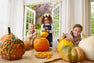Three smiling children carve pumpkins indoors, surrounded by various pumpkins and pumpkin seeds on a table. Bright daylight streams in through open double doors to a lush green garden outside, while one child shows off their Macy Drew Dress - Nantucket Navy Corduroy from The Beaufort Bonnet Company—a charming addition to any fall wardrobe.