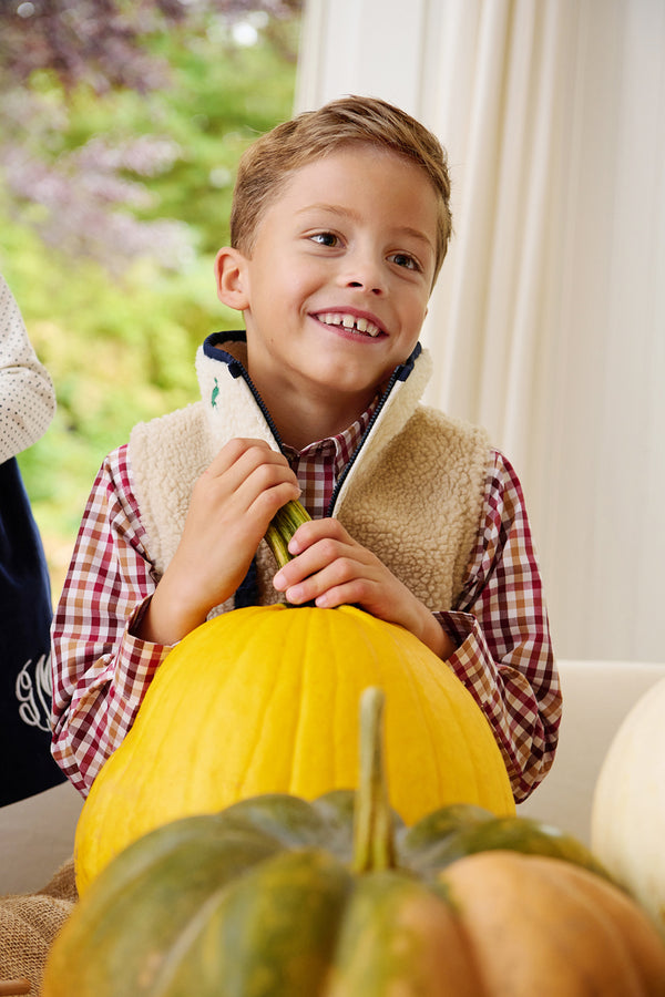 A smiling young boy wears The Beaufort Bonnet Company’s Dean’s List Dress Shirt in Easton Pond Plaid and a fleece vest as he holds a large yellow pumpkin, surrounded by other pumpkins. He is indoors, with greenery visible through a nearby window.