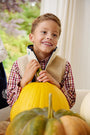 A smiling young boy wears The Beaufort Bonnet Company’s Dean’s List Dress Shirt in Easton Pond Plaid and a fleece vest as he holds a large yellow pumpkin, surrounded by other pumpkins. He is indoors, with greenery visible through a nearby window.