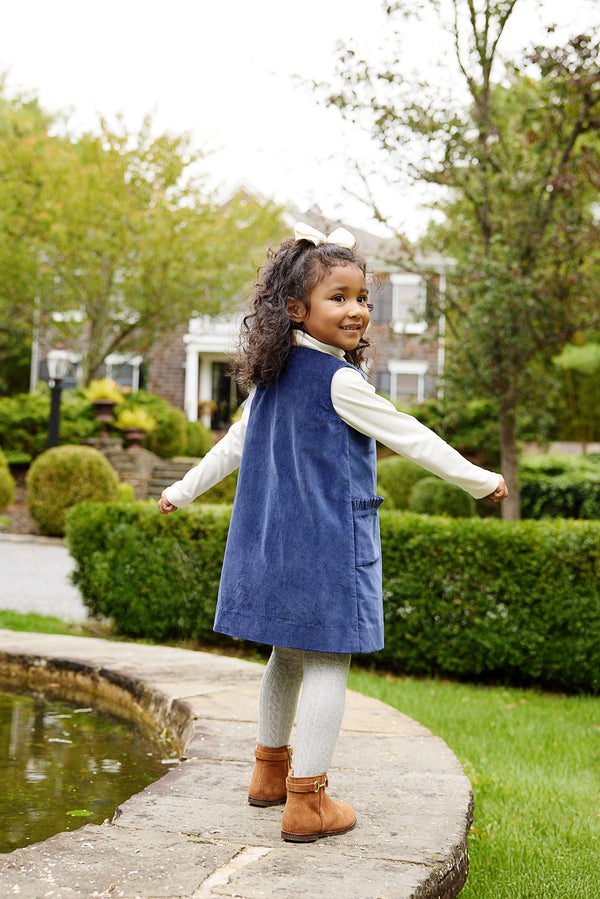 A young girl wears a Nantucket Navy Velveteen Jilly Jumper over a white long-sleeved shirt and light-colored tights while standing by a stone garden pond in a festive outfit.