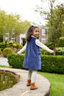 A young girl wears a Nantucket Navy Velveteen Jilly Jumper over a white long-sleeved shirt and light-colored tights while standing by a stone garden pond in a festive outfit.