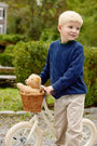 A young boy with blonde hair, wearing The Beaufort Bonnet Company’s Crawford Crewneck (Unisex) in Nantucket Navy and khaki pants, stands outside next to a white bicycle. A brown teddy bear sits in a wicker basket on the front of the bike, with green bushes and trees in the background.