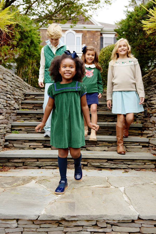 Four children in stylish outfits walk down outdoor stone steps, grinning. Leading the way, a girl wears The Beaufort Bonnet Company’s Short Sleeve Patsy’s Party Dress in Grier Green Velveteen with Nantucket Navy socks and shoes—an ideal holiday dress. Behind her, two girls and a boy are dressed in cozy sweaters paired with skirts or pants. Lush shrubs and a house complete the background.