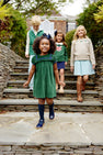 Four children in stylish outfits walk down outdoor stone steps, grinning. Leading the way, a girl wears The Beaufort Bonnet Company’s Short Sleeve Patsy’s Party Dress in Grier Green Velveteen with Nantucket Navy socks and shoes—an ideal holiday dress. Behind her, two girls and a boy are dressed in cozy sweaters paired with skirts or pants. Lush shrubs and a house complete the background.