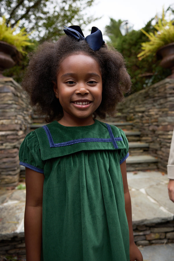 A young girl with curly hair, wearing The Beaufort Bonnet Company’s Short Sleeve Patsy’s Party Dress in Grier Green Velveteen with Nantucket Navy trim and bow, smiles outdoors in front of stone steps and greenery—a perfect choice for the holidays.