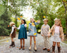 Five children stand on a gravel path in a wooded area, smiling and tossing autumn leaves into the air. They are dressed for fall in boots, sweaters, and the Long Sleeve Daphne Dress - Grier Green Herringbone by The Beaufort Bonnet Company. Behind them, green and yellow trees create a vibrant backdrop.